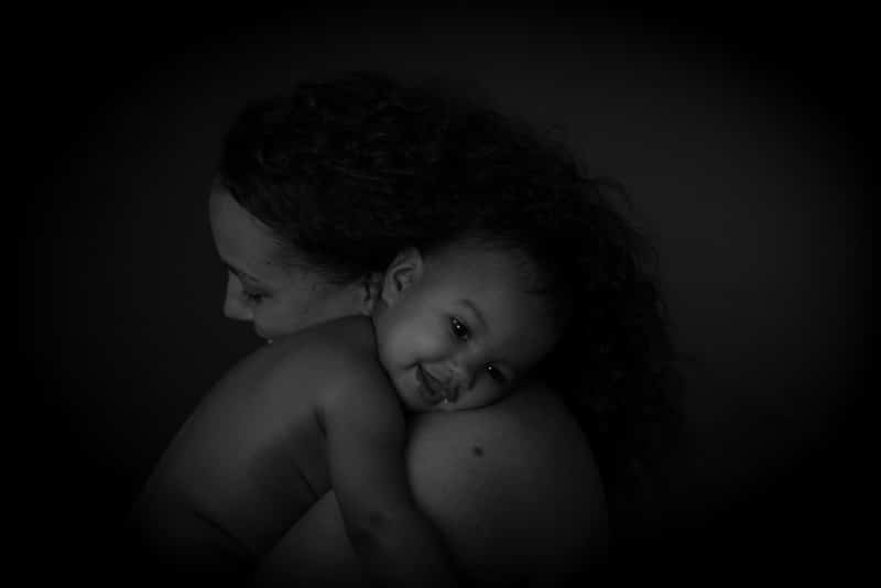 Black and white portrait of a baby smiling while cuddling into their mother’s shoulder during a professional baby photoshoot in Birmingham.