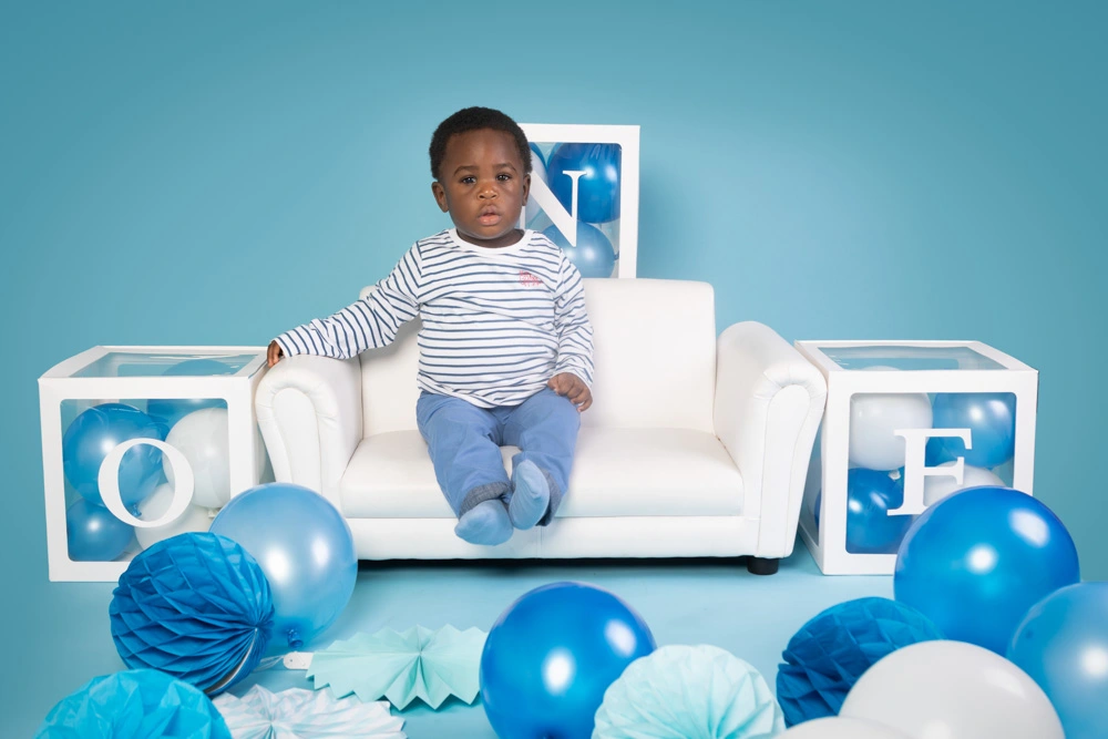 Young child sitting on a white sofa surrounded by blue balloons during a studio birthday photoshoot for children in Birmingham.