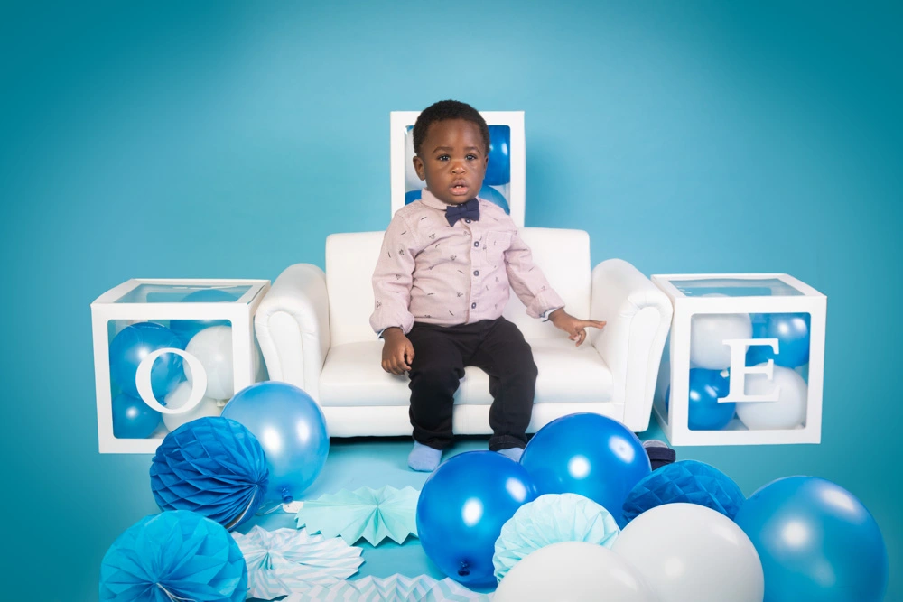 Young boy sitting on a white sofa surrounded by blue balloons during a studio birthday photoshoot for children in Birmingham.
