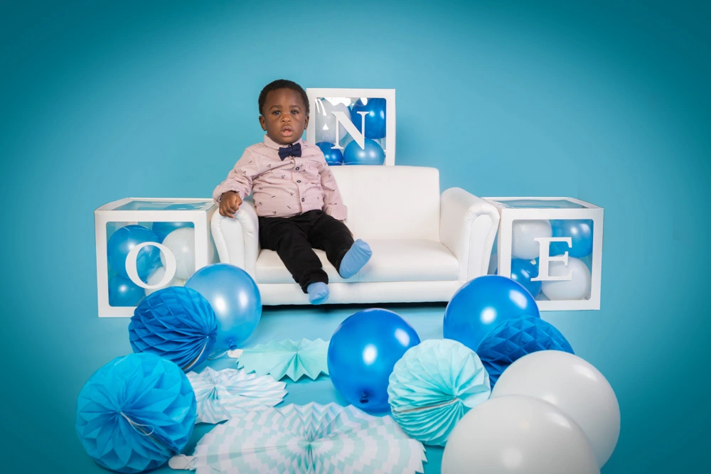 Young boy sitting on a white sofa surrounded by blue balloons and personalised blocks during a studio birthday photoshoot for children in Birmingham.