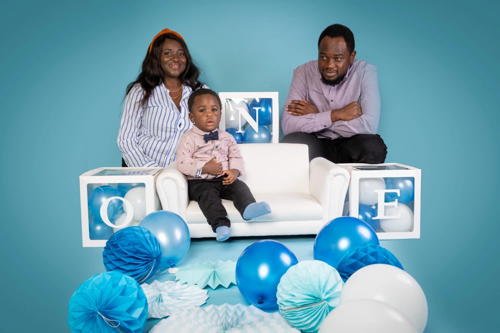 Parents posing with their young child seated on a white sofa surrounded by blue balloons during a studio birthday photoshoot for children in Birmingham.