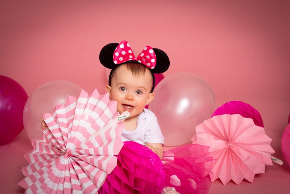 Baby girl wearing Minnie Mouse ears holding a pink fan during a studio birthday photoshoot for children in Birmingham.