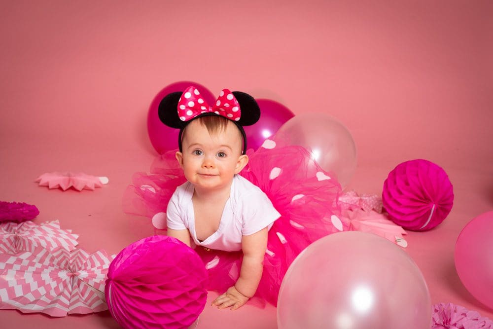 Baby girl wearing Minnie Mouse ears and a pink tutu crawling among balloons during a studio birthday photoshoot for children in Birmingham.