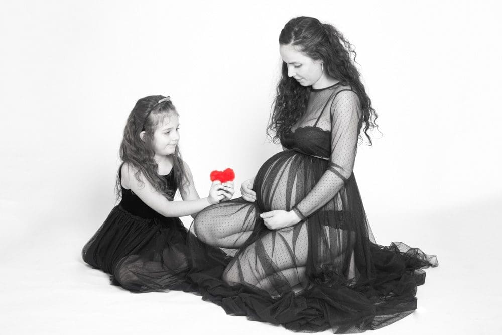 Pregnant mother kneeling in a flowing gown, looking down at her baby bump, while her young daughter sits beside her holding a small red heart.