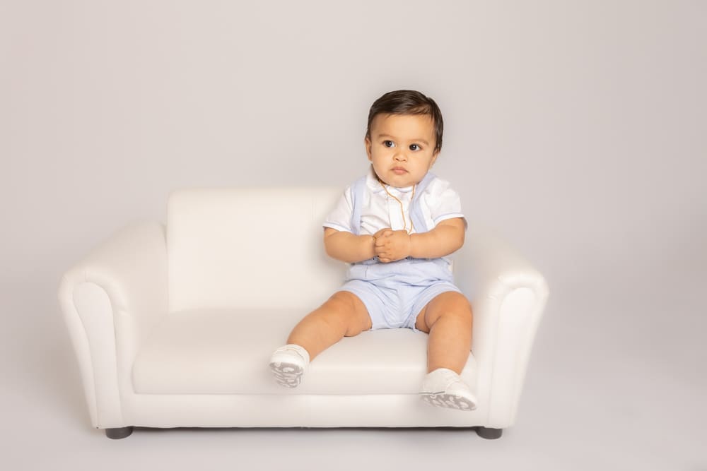 Baby sitting on a small white sofa during a professional baby photoshoot in Birmingham by an experienced baby photographer.