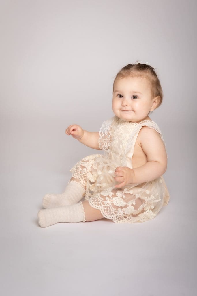 Baby girl sitting and smiling in a lace dress during a professional baby photoshoot in Birmingham.